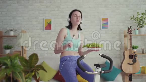 运动的年轻女子骑着一辆静止的自行车听音乐吃蔬菜视频的预览图