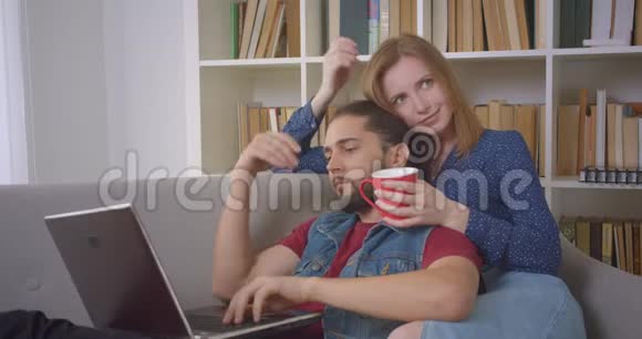 躺在女朋友身上的hipster家伙使用笔记本电脑当她在家温柔地抚摸他时她会和她交谈视频的预览图