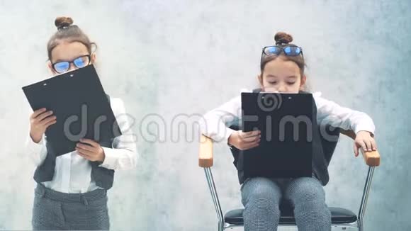 一个灰色背景的小商人在这期间女孩们拿着黑色的文件夹一个坐在椅子上这就是视频的预览图