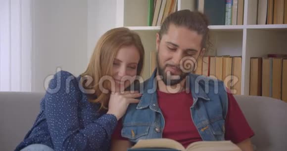 肖像镜头中年轻的白种人男女朋友坐在沙发上快乐地在家看书视频的预览图