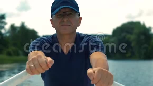 夏天一个瘦瘦的严肃的白发苍苍的男人戴着眼镜戴着蓝色的帽子穿着蓝色的t恤在河上划着一艘白色的船视频的预览图