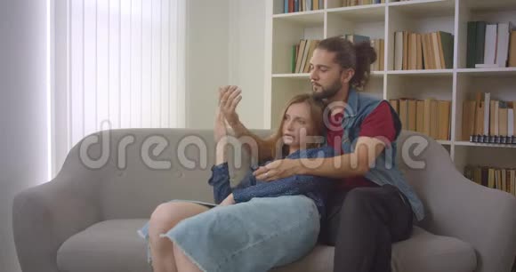 这张照片拍摄了年轻的白种人男女朋友坐在沙发上温柔地拥抱在家里视频的预览图