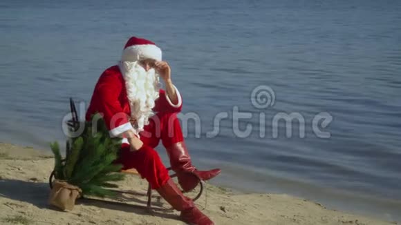 圣诞老人坐在沙滩上的雪橇上修理他的胡子眼镜和帽子视频的预览图