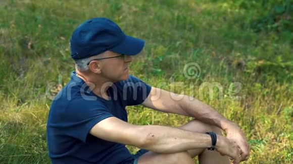一个夏天一个身材苗条头发严肃穿着蓝色t恤帽子和眼镜的人坐在森林里的绿草上视频的预览图