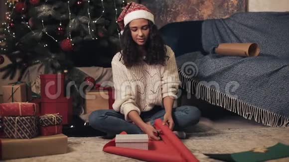 美丽幸福的女人戴着桑塔帽和白色套头衫准备礼物剪着包装纸坐在圣诞树下视频的预览图