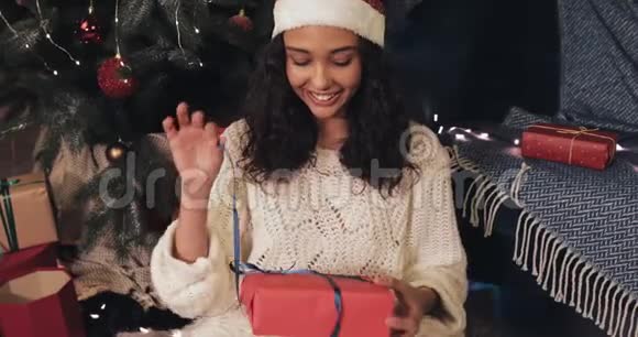 迷人的微笑女人坐在圣诞树下舒适的家庭背景打开红色礼物盒看上去视频的预览图
