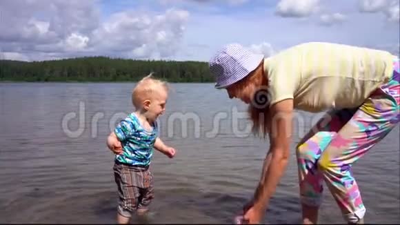 在晴朗的夏日在蓝天的映衬下妈妈和她的小儿子在一个森林湖的清澈的水中嬉戏玩耍视频的预览图