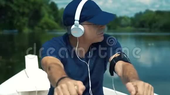 一个头发花白戴着白耳机戴着蓝帽子穿着t恤戴着眼镜的男子在白船上划桨视频的预览图