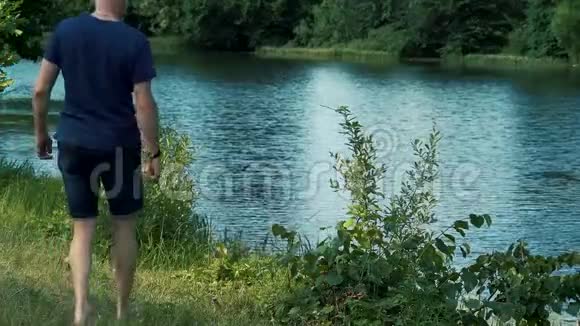 一个身披蓝色t恤帽子和眼镜的白发男子赤着脚走在河岸的绿草上视频的预览图