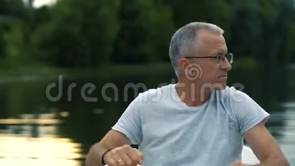 一个身材苗条头发灰白的严肃男子穿着灰色的t恤戴着眼镜在平静的河流上划着一艘白色的小船视频的预览图