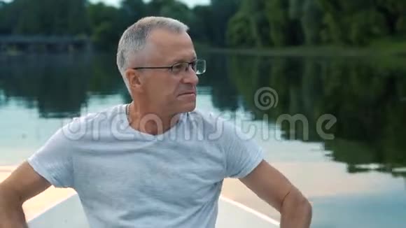 一个身材苗条头发灰白的严肃男子穿着灰色的t恤戴着眼镜在平静的河流上划着一艘白色的小船视频的预览图
