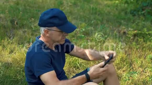 一个穿着蓝色t恤帽子和眼镜的头发花白的瘦长笑着坐在绿色的地上视频的预览图