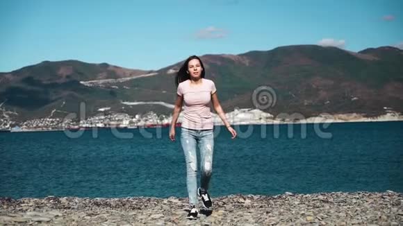 穿着粉红色衬衫和蓝色破牛仔裤的漂亮迷人的女孩或年轻女子在海和山上缓慢跳跃视频的预览图