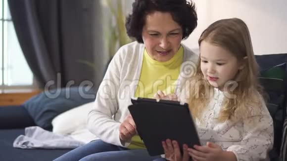 小女孩正在平板电脑上向高级女性展示新的有趣的网络游戏视频的预览图