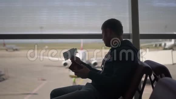 美国男子坐在机场的扶手椅上看着一张票目的是检查登机口视频的预览图