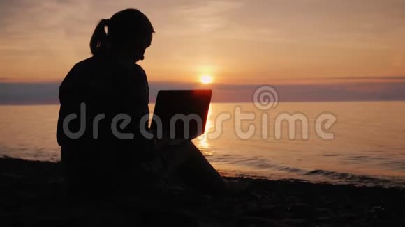 日落时分一位在海边拿着笔记本电脑工作的妇女的剪影视频的预览图