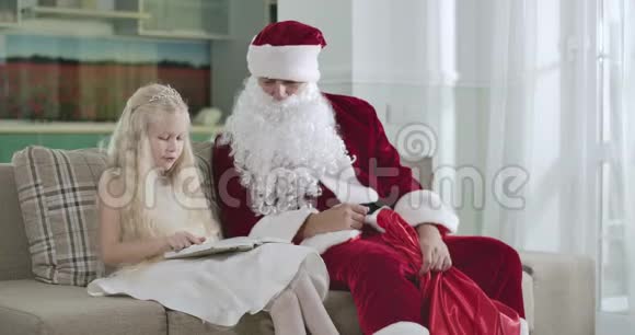 可爱的金发白种人女孩穿着优雅的白色连衣裙戴着皇冠坐在圣诞老人的马车上读着书视频的预览图