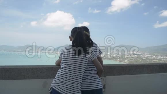 后景小女儿抱着妈妈开心地站着在观景台上观赏海湾的美景视频的预览图