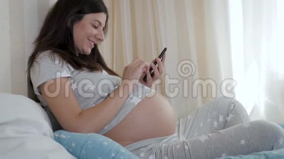 怀孕的白种人女人坐在床上使用电话和戳她的牙齿视频的预览图