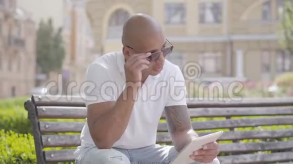 肖像英俊秃顶戴着太阳镜穿着白色t恤坐在长凳上打字的中东男子视频的预览图