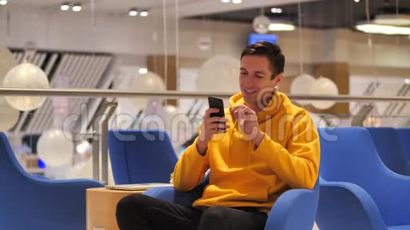 年轻的微笑快乐的男人穿着黄色的帽衫晚上在舒适的咖啡馆里使用智能手机视频的预览图