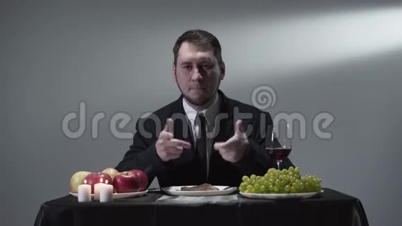 严肃的绅士穿着西装坐在小桌前安静地吃着肉葡萄苹果红酒和蜡烛视频的预览图