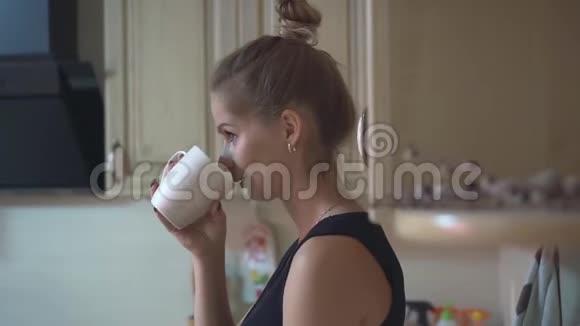 厨房里漂亮的女孩一个身穿黑色上衣的金发女郎在厨房里用白色杯子喝茶或咖啡视频的预览图
