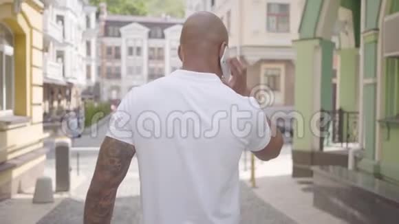 自信的中东秃子穿着白色t恤走在城市大街上用手机说话慢动作视频的预览图