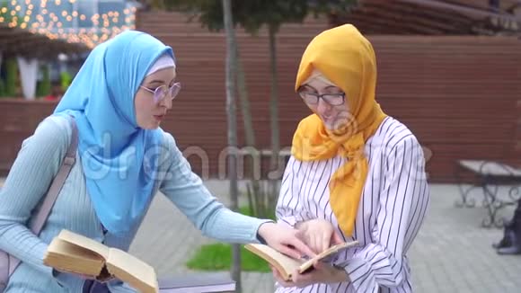两个戴着头巾戴着时髦眼镜带着笑容的穆斯林女孩坐在长凳上看书视频的预览图