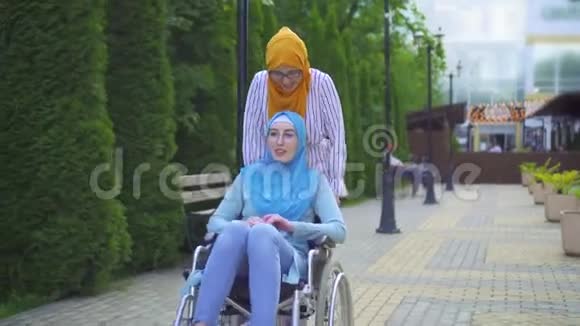 年轻的穆斯林残疾人戴着传统的围巾戴着轮椅的眼镜在街上和一个穆斯林朋友玩视频的预览图