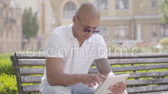 肖像英俊秃顶的中东男子戴着太阳镜和白色t恤平板电脑坐在长凳上视频的预览图