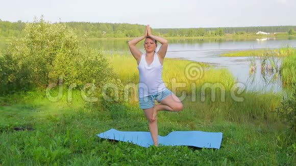 早晨一位黑发女子在湖岸树上摆姿势练习瑜伽视频的预览图