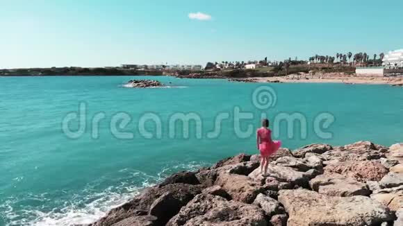 穿红色衣服的女人走在岩石海滩上举起双手分开在空中看着蓝色的海洋和天空风在拍打她的裙子视频的预览图