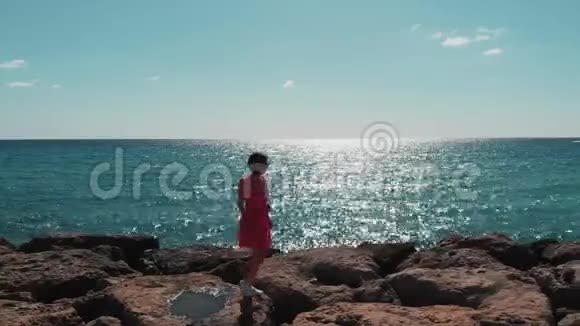 穿着红色裙子的可爱女士在海上码头的岩石上随风拍打蓝色海水上有阳光反射天气晴朗炎热视频的预览图