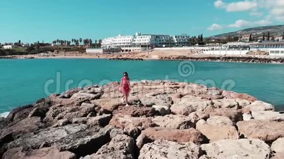 穿着红色漂亮裙子的女人独自走在塞浦路斯珊瑚湾的岩石码头上背景是酒店和海滩蓝色的海波视频的预览图