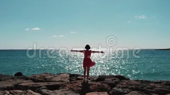 穿着红色连衣裙的女人的剪影手臂分开在空中站在岩石海滩码头上迎着阳光风拍打着她的裙子吉吉视频的预览图