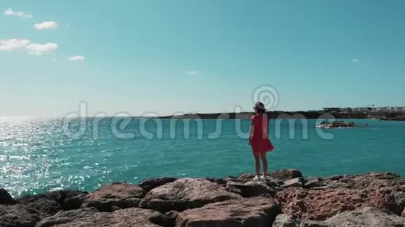 迷人的女人穿着红色的衣服站在地中海的岩石悬崖上看着蓝色的海水和天空gi的无人视角视频的预览图