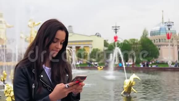 可爱的女孩微笑着在智能手机上键入一条信息时髦的衣服黑色夹克视频的预览图