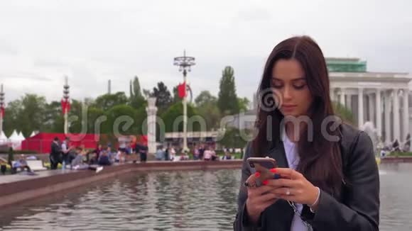 公园里迷人的微笑的女孩坐在喷泉旁用智能手机视频的预览图