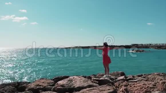 穿着红色裙子的女人双手分开迎着阳光站在岩石悬崖码头享受着风和阳光海浪撞击岩石视频的预览图