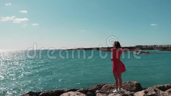 在阳光明媚的夏日塞浦路斯珊瑚湾穿着红色衣服的女士双手分开站在岩石海滩悬崖码头上招展Ae视频的预览图