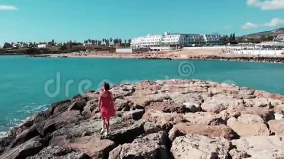 可爱的女性穿着红色的裙子独自走在岩石海滩码头享受阳光和热风波浪与岩石碰撞阳光反射视频的预览图