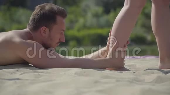 迷人的男人躺在沙滩上抱着试图近距离行走的年轻女人的腿户外训练积极主动视频的预览图
