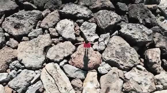 穿红色衣服的女人双手分开站在岩石上俯视着拍摄戴着红裙子的女士站在岩石灯塔上视频的预览图