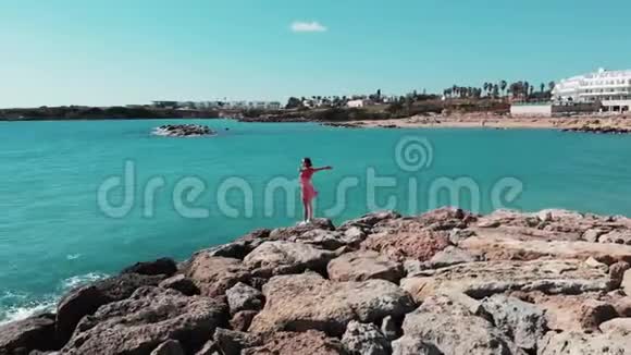 双手分开的女人穿着红色可爱的裙子风拍打着站在岩石悬崖上看着太阳路和蓝色的海水视频的预览图