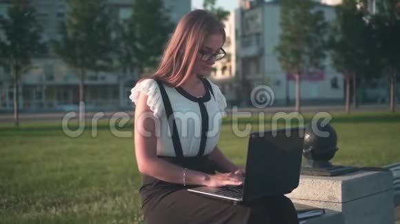 商务女性穿着白色衬衫和眼镜坐在长凳上在笔记本电脑上工作这个女孩对这项工作很满意视频的预览图