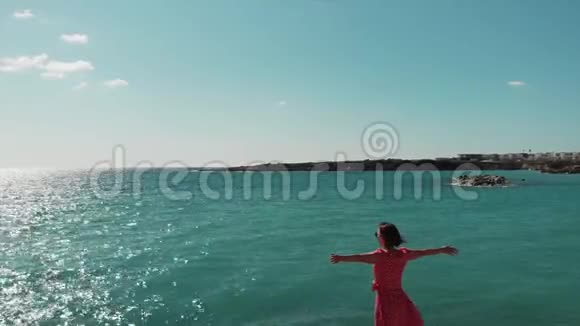 可爱的女人穿着红色连衣裙双手分开站在悬崖上穿红色衣服的女士在岩石海滩上手臂分开在空中空中射击视频的预览图