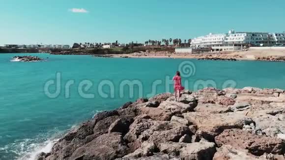 在岩石海滩悬崖上穿红色衣服的女人穿红色裙子的女士在塞浦路斯珊瑚湾享受阳光和温暖的风成功的女性视频的预览图