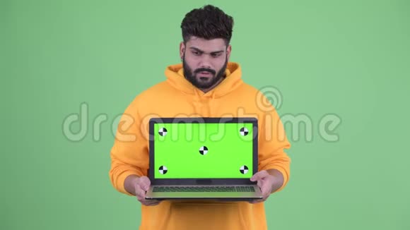 快乐的年轻体重超重留着胡子的印度男人一边看着笔记本电脑一边思考视频的预览图