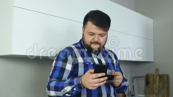 一个胖子站在厨房里看着电话喝着咖啡一个大男人在厨房里盯着视频的预览图